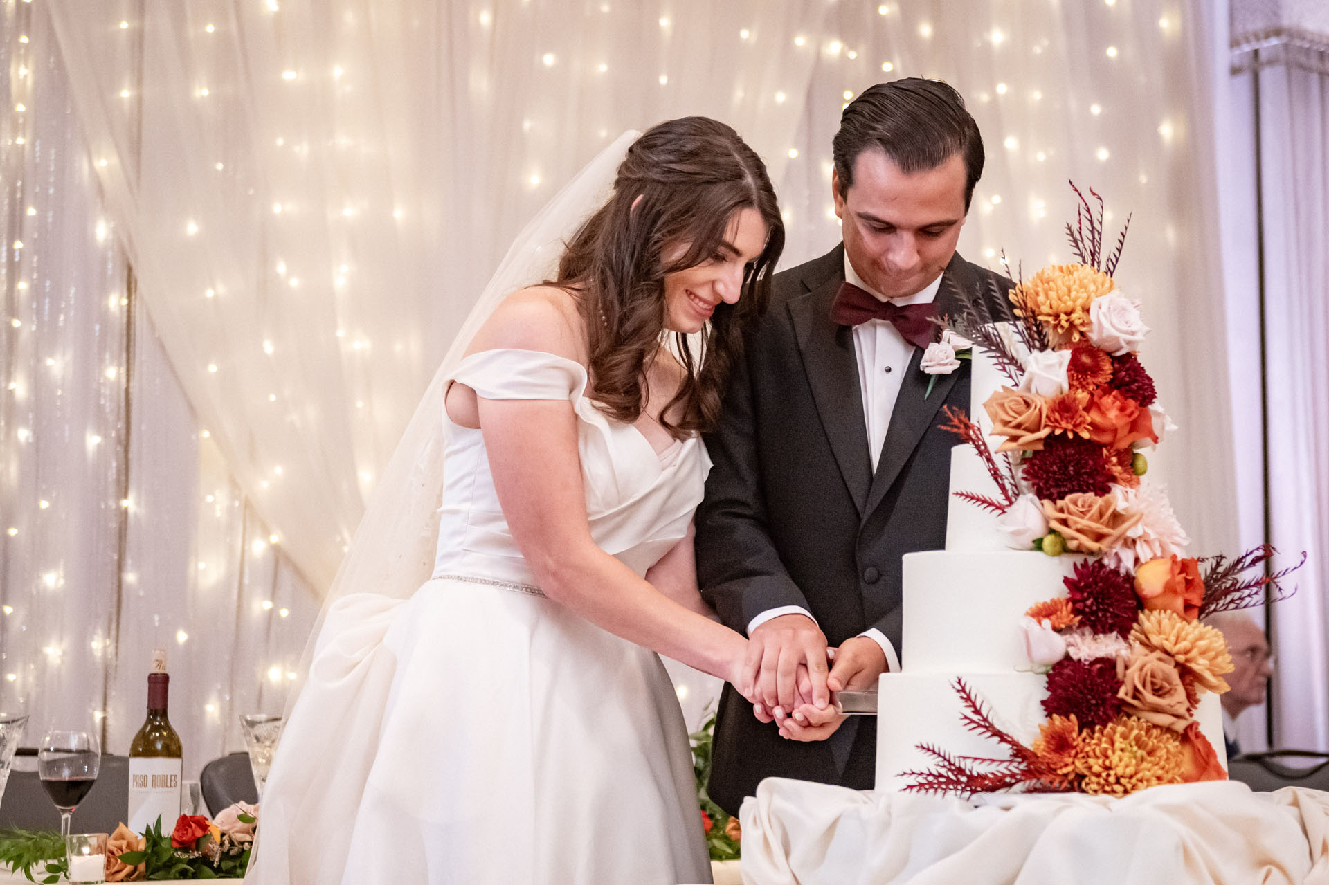 Smiling bride and groom cut autumn-themed wedding cake in front of sparkling backdrop 