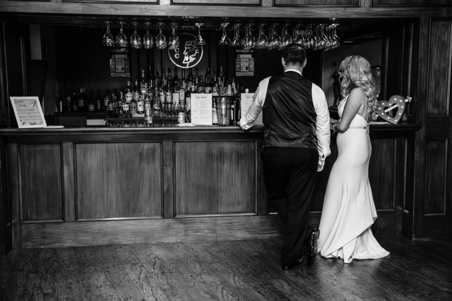 Classic, black-and-white candid portrait of bride and groom in front of large, open bar area